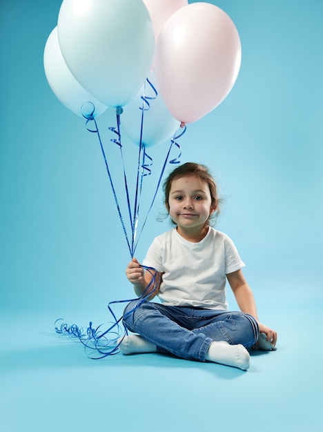 Cute little girl sitting on blue surface with soft shadow and posing to front with balloons in the hand