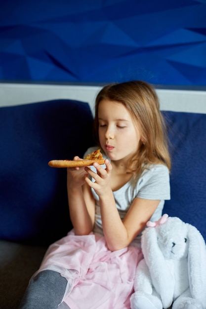 Cute little girl sitting on couch and eating piece of italian Pizza at home, yummy meal