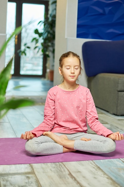 Cute little girl sitting on fitness mat in a lotus position while doing yoga at hom