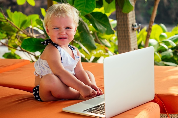 A cute little girl sitting on orange lounger with a laptop on the beach on a warm summer day. holiday on a seashore tropical green park background concept.fair-haired business kid one year old