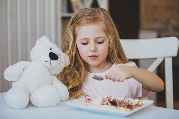 Cute little girl sitting at a table in a cafe and feed a favorite toy Bear ice cream