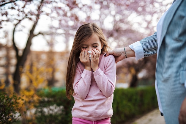 Cute little girl sneezing in park while she is walking in park with her mother. Allergy, flu, virus concept.