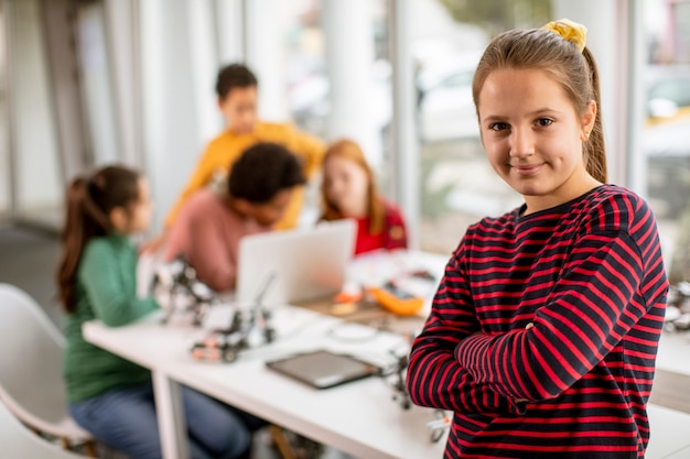 Cute little girl standing in front of  group of kids programming electric toys and robots at robotics classroom