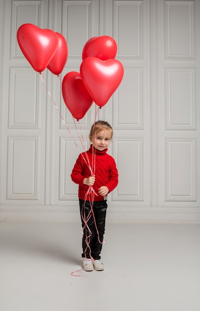 Cute little girl stands and holds with red heart shaped balloons on white