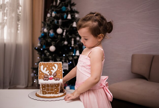 A cute little girl stands near the table and looks at the gingerbread house