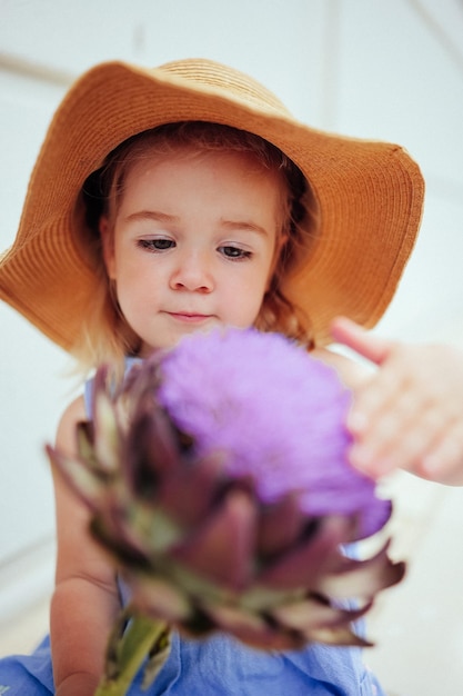 Cute little girl in straw hat touching big artichoke flower