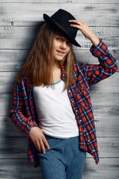 Cute little girl in stylish hat standing at the wall in studio. kid isolated on wooden background, child emotion, schoolgirl photo session