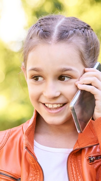 Cute little girl talking Hold phone near face Young woman smile Not looking at camera Outdoor school Green background Smiling speach Happy expression Student lifestyle Vertical