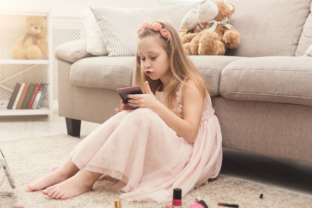 Cute little girl trying her mom's cosmetics. Pretty kid looking at mirror, sitting on floor carpet among lots of beauty products. Small fashionista doing make up, copy space