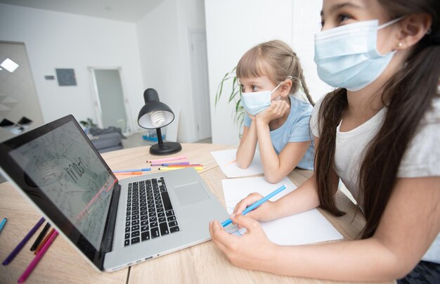 Cute little girl wearing a medical mask on her face doing distance learning using a laptop at home in the bedroom. A girl in a protective mask on her face holds her head in her hand. Distance Learning