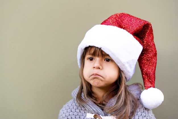 Bambina sveglia che indossa il cappello di babbo natale contro uno sfondo verde della parete all'aperto nel periodo natalizio bambina imbronciata per il regalo di natale faccia buffa smorfia infanzia e vacanze di natale