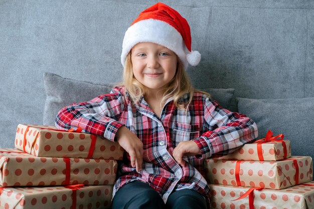 Cute little girl wearing Santa Claus hat plaid woolen pajama sitting on couch between present box