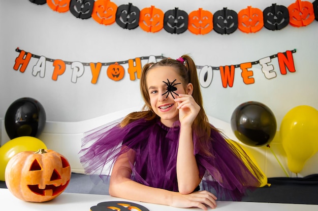 Bambina carina che indossa un cappello da strega seduta dietro il tavolo nella stanza decorata a tema di halloween covid19