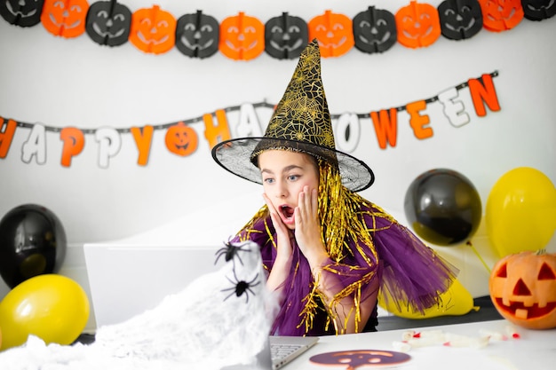 Cute little girl wearing witch hat sitting behind table in Halloween theme decorated room Covid19