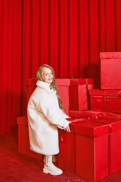 Cute little girl in white coat by the huge red christmas\
presents boxes on red background. holidays