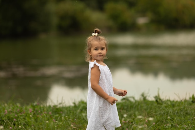 A cute little girl in a white dress stands in the grass on the shore of a lake on a summer evening Sunset at the lake