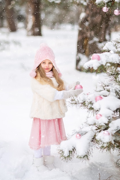 Cute little girl in a white fur coat and pink fluffy skirt in the forest in winter dresses up the Christmas tree. Pink Christmas toys.