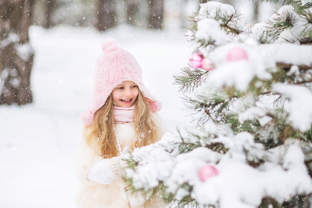 Cute little girl in a white fur coat and pink fluffy skirt in the forest in winter dresses up the Christmas tree. Pink Christmas toys.