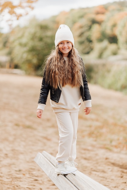 Милая маленькая девочка в белой шляпе и черной кожаной куртке дурачится в осеннем парке
