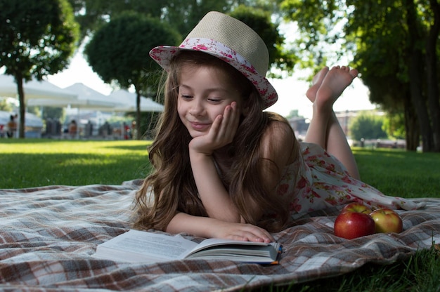 Cute little girl in the wicker hat and dress with flowers reading a book on a sunny summer day