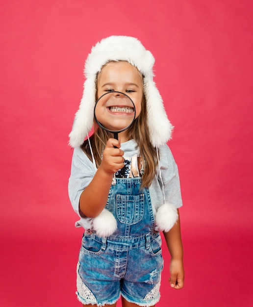 Cute little girl in a winter hat looking through a magnifying glass on a pink background.