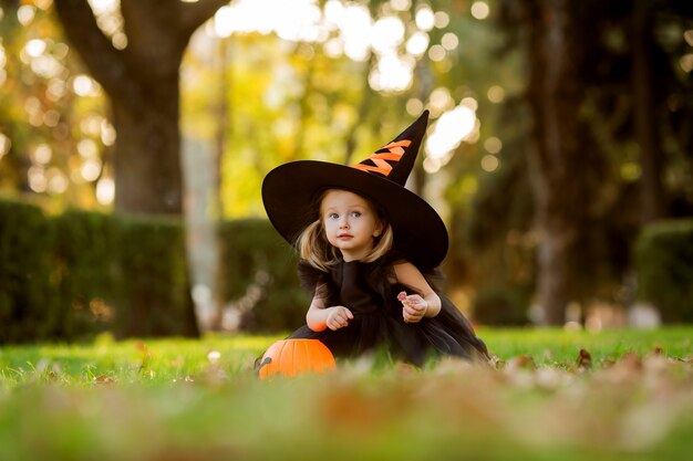 A cute little girl in a witch costume walks in the street with a candy in the shape of a pumpkin. 