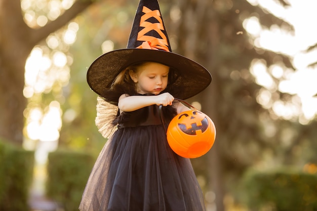 A cute little girl in a witch costume walks in the street with a candy in the shape of a pumpkin. 