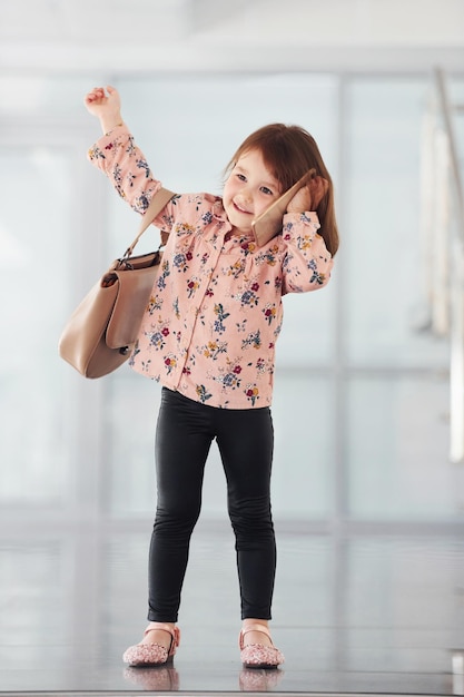 Милая маленькая девочка с сумкой в помещении в офисе или аэропорту разговаривает по телефону