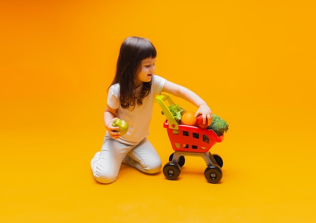 黄色のbackgroundstudio写真で隔離のスーパーマーケットからの食料品のバスケットを持つかわいい女の子