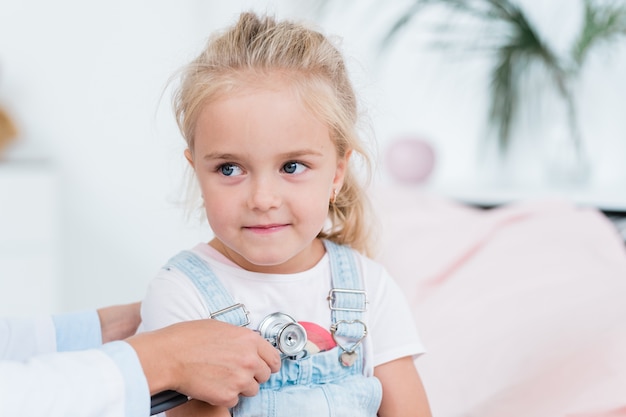 Cute little girl with blond hair looking at doctor using stethoscope during medical treatment in hospital