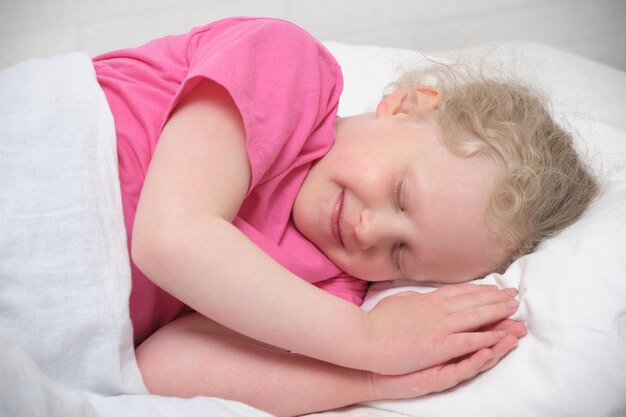 Cute little girl with blond hair and a pink tshirt sleeps on a white bed