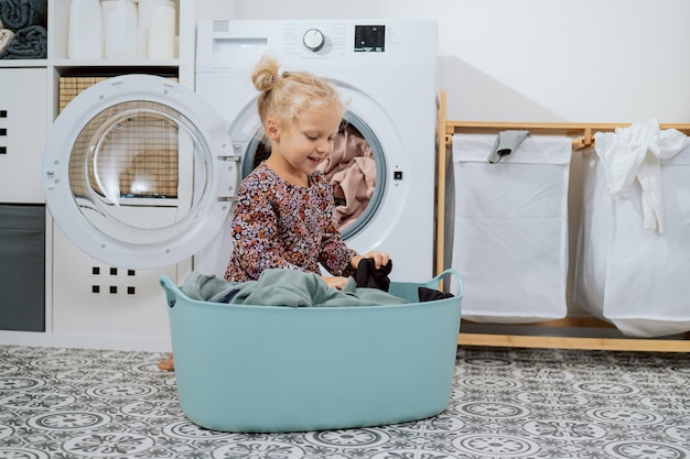 Cute little girl with blonde hair small child sits in bathroom\
laundry room by large bowl full of clothes daughter throws things\
into washing machine helps mom with daily chores