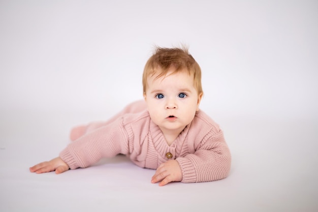 A cute little girl with blue eyes in a pink knitted jumpsuit is lying on her stomach looking at the camera on a white background isolated