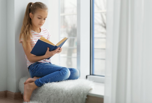 Милая маленькая девочка с книгой сидит дома на подоконнике