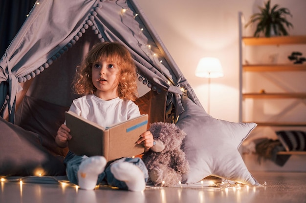 Cute little girl with curly hair is in the tent in domestic\
room with interesting book