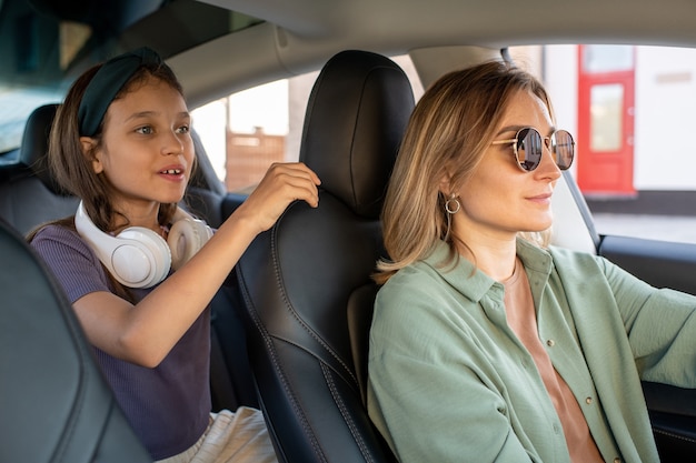 Bambina sveglia con le cuffie che tengono dal sedile di sua madre che guida l'auto