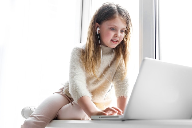 Cute little girl with headphones and a laptop is sitting at home on the windowsill