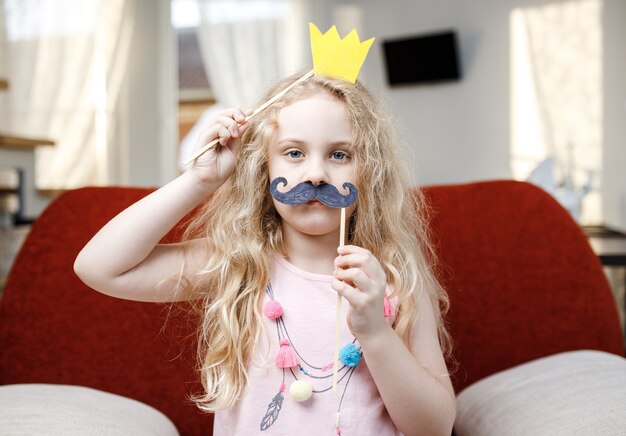 Cute little girl with paper crown and mustaches while sitting on red chair at home.