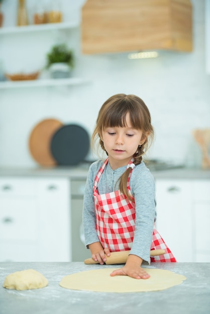 格子縞のエプロンにピグテールを付けたかわいい女の子が、ピザ生地を自分でこねます。子供のための料理教室。