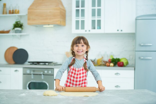 격자 무늬 앞치마에 땋은 머리를 가진 귀여운 소녀가 피자 반죽을 직접 반죽합니다. 어린이를위한 요리 교실.