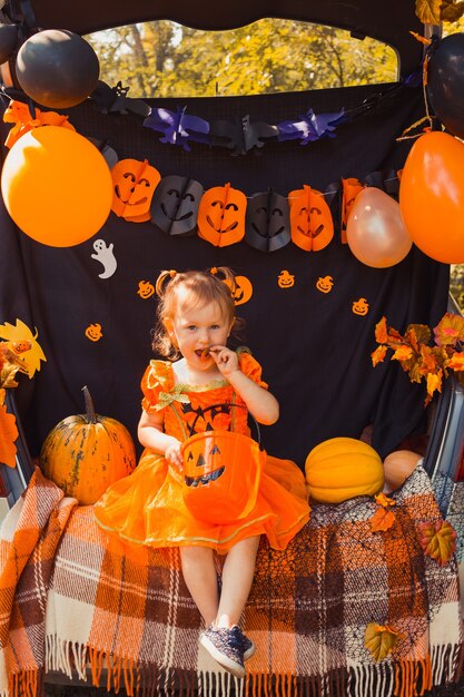 Cute little girl with a pumpkins eating candy from buckets sitting on trunk of car