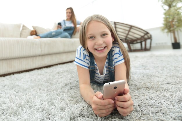 コピースペースのあるリビングルームの写真のカーペットの上に横たわっているスマートフォンを持つかわいい女の子