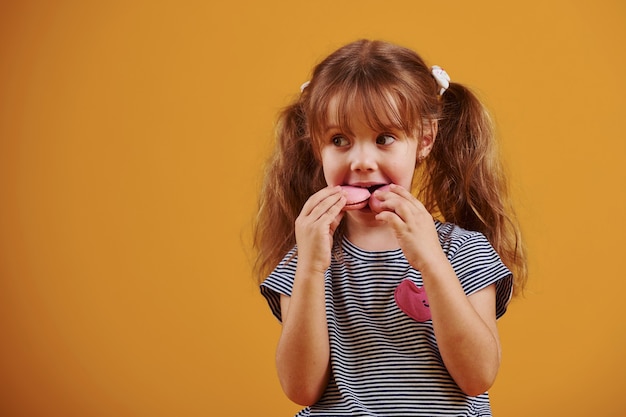 Милая маленькая девочка со сладкой едой в студии на желтом фоне.