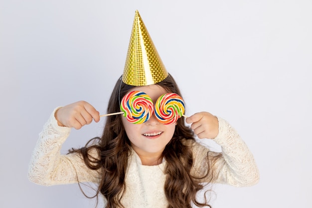 Bambina sveglia con due grandi lecca-lecca sugli occhi in un cappello festivo su sfondo bianco isolato con una bandierina. spazio per il testo. la bambina festeggia il compleanno, il concetto di vacanza