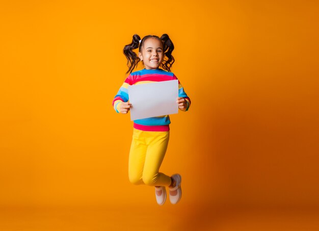 Cute little girl with a white sheet of paper. yellow background little girl holding blank sheet of paper