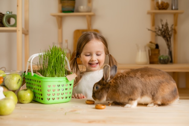 家の木製のキッチンでかわいい女の子はウサギの新鮮な草を養います