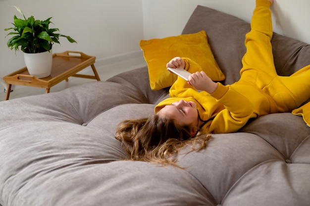 cute little girl in yellow clothes is lying on her back on bed and holding a smartphone in her hands