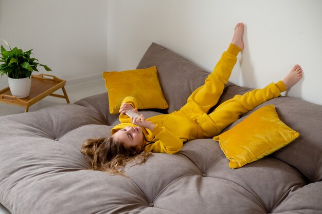 cute little girl in yellow clothes is lying on her back on bed and holding a smartphone in her hands