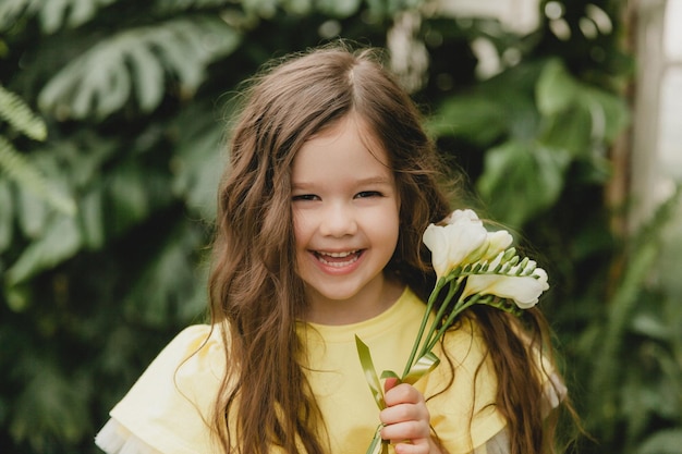 Cute little girl in a yellow dress holding spring flowers in her hands standing against the background of leaves