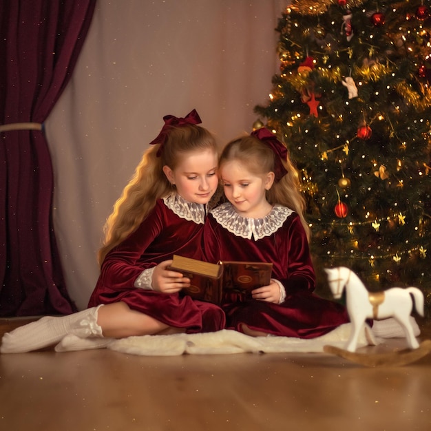 Cute little girls in christmas clothes are reading a book on the background of a christmas tree